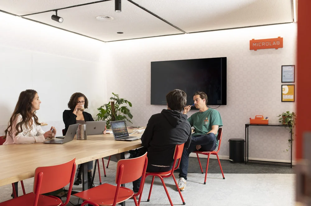 Vier mensen zitten in een vergaderruimte aan de Aert van Nesstraat bij Microlab, in gesprek rond een tafel met laptops.